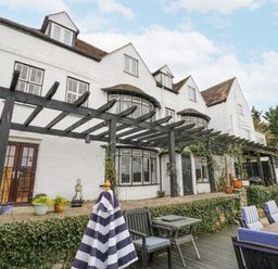 An exterior view of a white house with multiple windows and dormers a black pergola with climbing plants a patio with potted plants and wooden outdoor furniture with striped cushions and a folded striped umbrella at Riverside Manor in Evesham