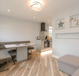 A dining area with grey bench seating and a white table next to a kitchen with stainless steel appliances and wooden flooring at Number 9 in California Norfolk
