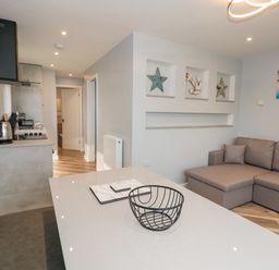 A living area with a gray sofa and a table with a wire basket and placemats in a kitchen space at Number 9 California Norfolk