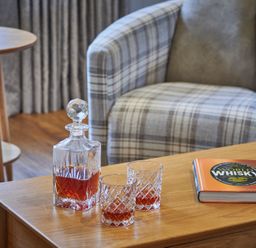 A wooden table with a crystal decanter and two glasses containing whisky and a book titled World Whisky next to a plaid armchair at The Filly in Humshaugh