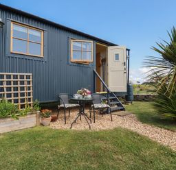 A small blue corrugated metal building with two windows and an open door next to a small patio with a round table and two chairs on gravel at Dot May Porteath near Polzeath