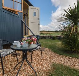 A small outdoor seating area with a table and two chairs outside a building with open door and window at Dot May Porteath near Polzeath