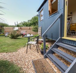 An outdoor garden area with a small table and two chairs next to stairs leading to a blue cabin with an open door at Dot May Porteath near Polzeath