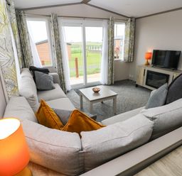 A living room with a corner sofa a small coffee table a TV on a cabinet and windows with curtains at Sandy Bay Retreat in Hasguard Cross near Broad Haven