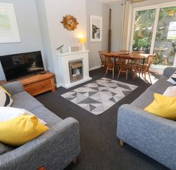 A living room with two grey sofas with pillows a geometric rug a wooden dining table with chairs a fireplace a flat screen TV and a sliding glass door at 8 Rosewall Cottages in St Ives