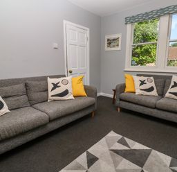 A living room with two gray sofas with decorative pillows a geometric patterned rug and a window at 8 Rosewall Cottages in St Ives