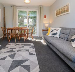 A living room with a gray sofa with cushions a patterned rug a wooden dining table with chairs and sliding glass doors at 8 Rosewall Cottages in St Ives