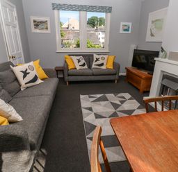 A living room with two gray sofas with yellow and patterned cushions a geometric rug a wooden dining table chairs a fireplace and a television at 8 Rosewall Cottages in St Ives