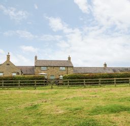 A stone house with chimneys behind a wooden fence and hedge with a grassy field in front at The Granary in Longwitton near Morpeth