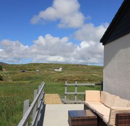 An outdoor patio with a cushioned sofa and table overlooking grassy fields and hills with wind turbines at Eala Bhan Cottage in Clachan near Balivanich
