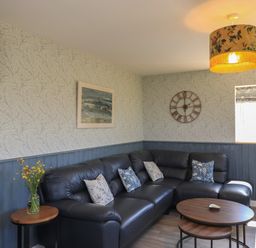 A living room with a black sectional sofa pillows round wooden coffee tables a wall clock a wall painting and a window at Eala Bhan Cottage Clachan near Balivanich