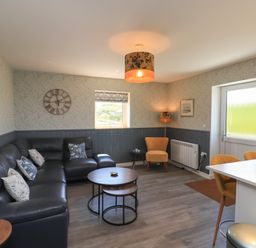 A living room with a black sectional sofa and cushions a round wooden coffee table a yellow armchair a wall clock and a door at Eala Bhan Cottage in Clachan near Balivanich