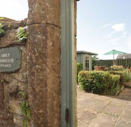 A stone entrance wall with a sign reading Abbey Farmhouse Entrance and a garden patio with chairs and a green umbrella at Abbey Farm House in St Bees