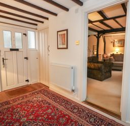 An entryway with a patterned rug a white panel door a radiator and a view into a living room with sofas and exposed ceiling beams at Poppy Cottage in Beaminster