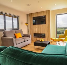 A living room with two sofas a yellow armchair and ottoman a TV mounted on wood paneling and a coffee table with fruit at Mynydd Y Glyn Lodge in Llanfyllin near Llanrhaeadr-Ym-Mochnant
