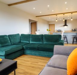A living room with a green sectional sofa, a gray sofa with orange cushion, and a kitchen area with bar stools and hanging lights at Mynydd Y Glyn Lodge in Llanfyllin near Llanrhaeadr-Ym-Mochnant