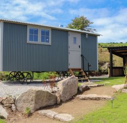A small corrugated metal shepherds hut on wheels with a white door and window beside a gravel path and large rocks at Bosulla Shepherds Hut in Penzance