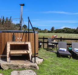An outdoor area with a wooden hot tub with steps two lounge chairs with towels a small table with flowers and chairs in a grassy fenced field at Bosulla Shepherds Hut in Penzance