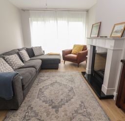 A living room with a grey sectional sofa with cushions a brown armchair a fireplace with framed art and a wooden cabinet at 19A Lighthouse Village in Fenit County Kerry