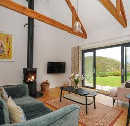 A living room with a green sofa a black coffee table a pink armchair a wood burner and large sliding glass doors showing a hill outside at Pendower Cottage in Porthgwarra