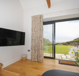 A room with a wall-mounted TV a basket of firewood patterned curtains sliding glass doors leading to a garden and flowers on a table at Pendower Cottage in Porthgwarra