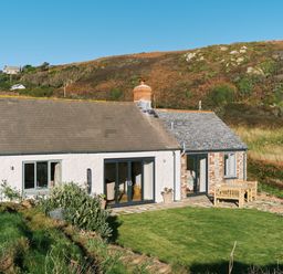 A cottage with white and brick walls with a lawn and outdoor wooden furniture at Pendower Cottage in Porthgwarra