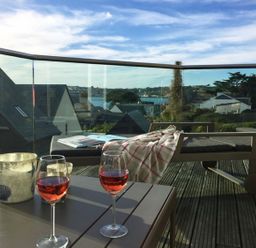 A balcony with two glasses of rose wine on a table and a lounge chair with a blanket overlooking rooftops and water at Gull Rock 4 in Rock