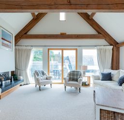 A living room with white sofas, armchairs, a wooden coffee table, large windows, exposed wooden beams, and a fireplace at Gull Rock 4 in Rock