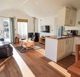 A living room with a leather sofa wooden coffee table and fireplace next to a kitchen with white cabinets and wooden countertops at Pippin Lodge in Allithwaite near Cartmel