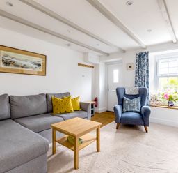 A living room with a gray sofa yellow pillows a blue armchair a wooden coffee table and a window seat with cushions and flowers at Stokes Cottage in St Minver
