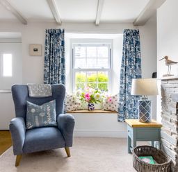 A living room with a blue armchair a window seat with cushions and flowers a side table with lamp and a stone fireplace at Stokes Cottage in St Minver