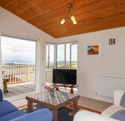 A living room with a wooden ceiling a coffee table with flowers a blue armchair a white sofa a television and large windows with a view at Tamar View Lodge Millbrook