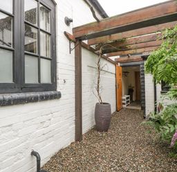 A gravel pathway with a large planter under a wooden pergola beside a white brick wall with black framed windows at The Old Bakehouse in Pembridge