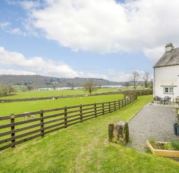 A grassy yard with wooden fence and outdoor seating next to a stone and white house overlooking fields and a lake at Shepherds Cottage in Hawkshead