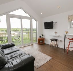 A living room with a black leather sofa a wall mounted TV a small wooden nesting table and stools near large windows overlooking a balcony with chairs at Mallard Lodge in Moscow near Galston