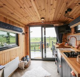 A kitchen with wooden walls and ceiling a countertop with a sink stove and cutting board and sliding glass doors showing a green landscape at Greenacres Carriage in Maybole