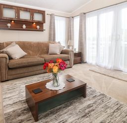A living room with a brown sofa and pillows a wooden coffee table with flowers a side table with a lamp and curtains at Gwel Eryri in Penmynydd near Llanfairpwllgwyngyll
