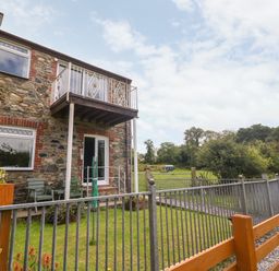 A stone house with a small balcony and garden area enclosed by metal and wooden fences at Coed y Glyn Bach Equestrian in Penygroes