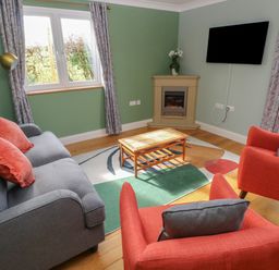 A living room with a gray sofa and orange chairs a wooden coffee table a wall-mounted TV and a corner fireplace at The Annexe Meadow Croft in Haverfordwest