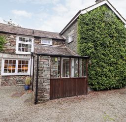 Exterior view of a stone house with white framed windows and a porch covered in glass at Holly Cottage in Grasmere