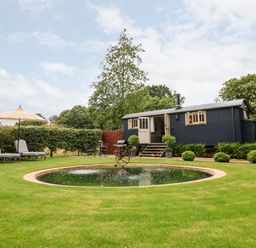 A garden with a circular pond lounge chairs with an umbrella and a dark blue shepherds hut at Utopia in Metfield near Harleston