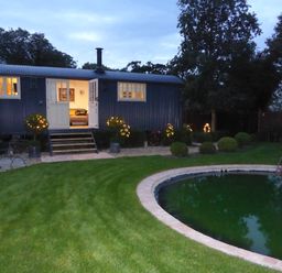 A garden with a grass lawn a circular pond and a small building with open doors and lit windows at Utopia in Metfield near Harleston