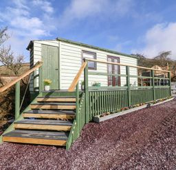 A small wooden cabin with stairs and a railing on a gravel surface surrounded by shrubs and hills at Nyth Y Bioden (Magpie's Nest) in Llanaelhaearn near Trefor