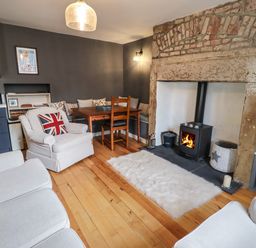 A living room with a wood burning stove a wooden dining table with chairs and light-colored armchairs at Grebe Cottage in Alnmouth