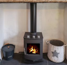 A black wood-burning stove with fire inside placed in a white plastered fireplace with a black ash bucket and a white woven basket at Grebe Cottage in Alnmouth