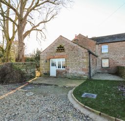 A stone cottage with a white door and windows surrounded by gravel driveway grass and a tree at Luham Cottage Edenhall near Langwathby