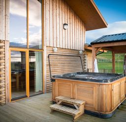 An outdoor wooden hot tub on a deck next to a cabin with large windows at Aurae in Cawdor near Inverness