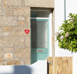 A stone wall with a green door and a red heart house number plate 11 with a wooden gate and plants at 11 Belgravia Street in Penzance