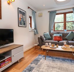 A living room with a gray sofa and coffee table with breakfast items a television on a wooden cabinet and blue curtains at Cherry Tree Cottage in Windermere