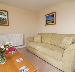 A living room with a beige sofa with two cushions two framed pictures on the wall and a wooden coffee table with a vase of flowers at Hunstone Barn in South Molton